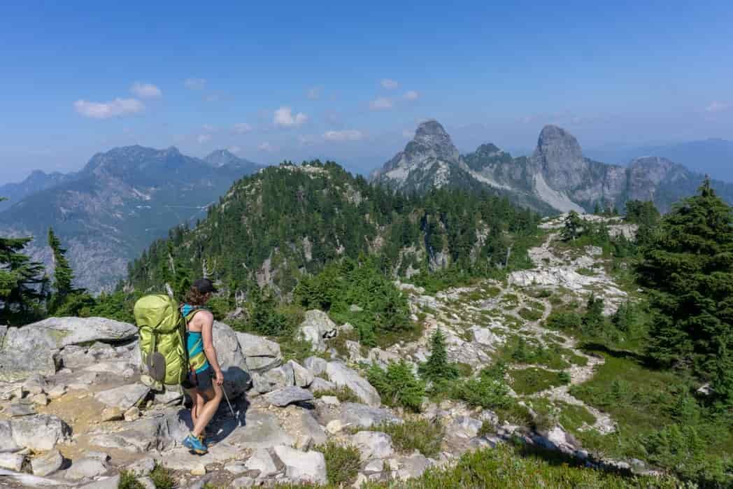 A woman wearing a large backpack walks along the Howe Sound Crest Trail with the Lions in the background.