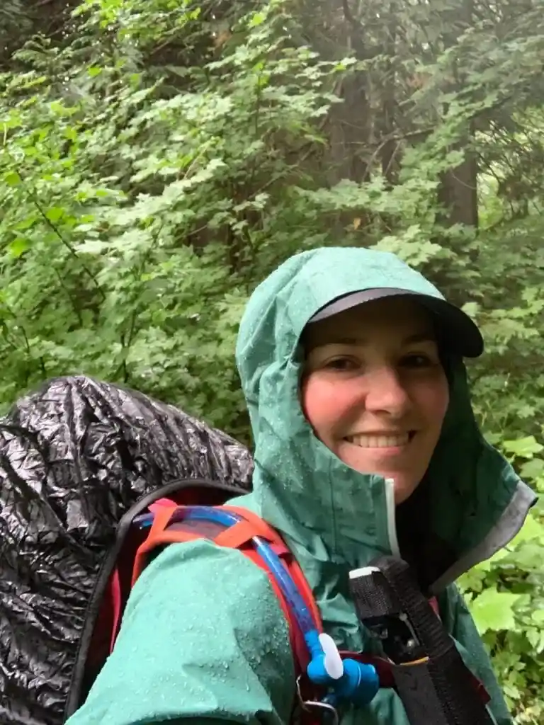 A hiker takes a selfie in the rain. She is wearing a rain jacket, a base ball camp, and a backpack with a rain cover on it.