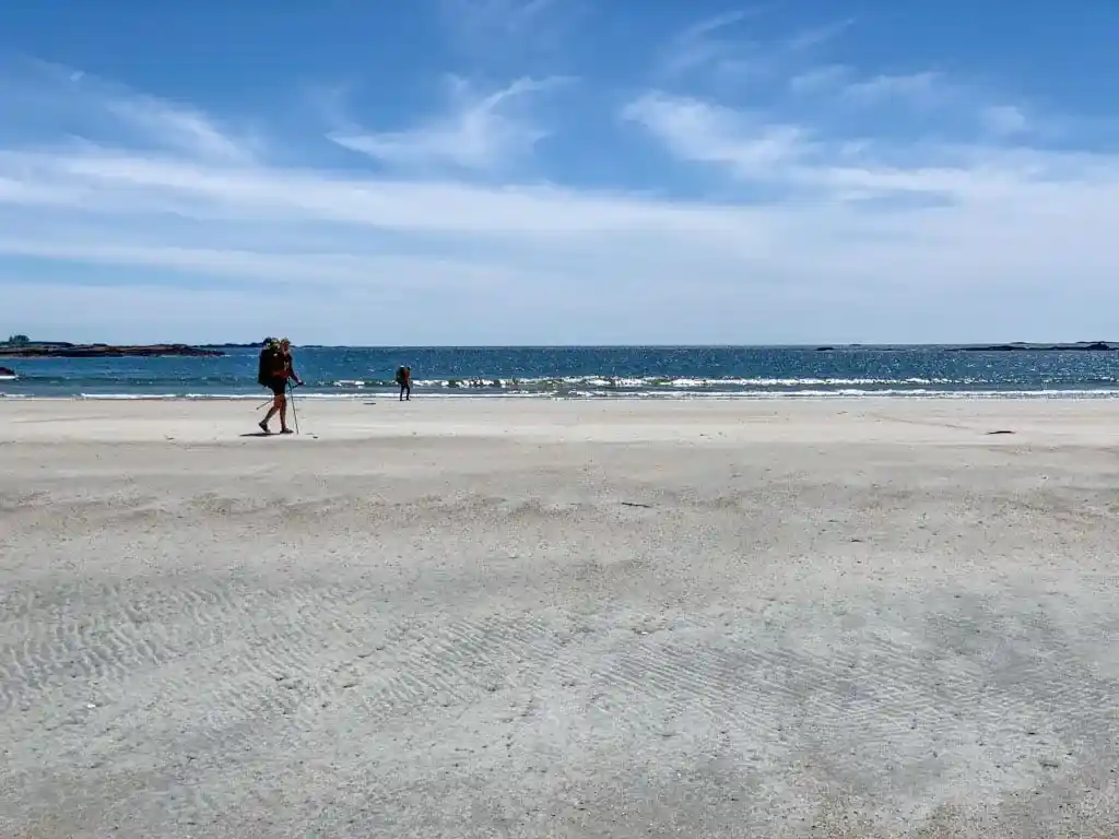 Two hikers walk across a broad sandy beach near Sandpiper Creek on the Tatchu Trail