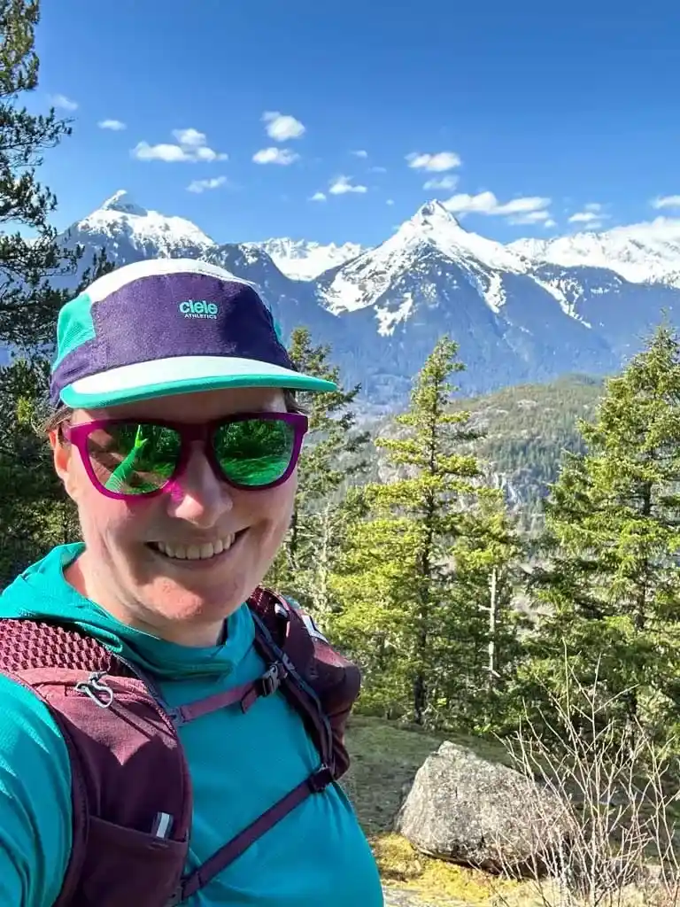 A woman wearing a Ciele trail running cap takes a selfie in the mountains