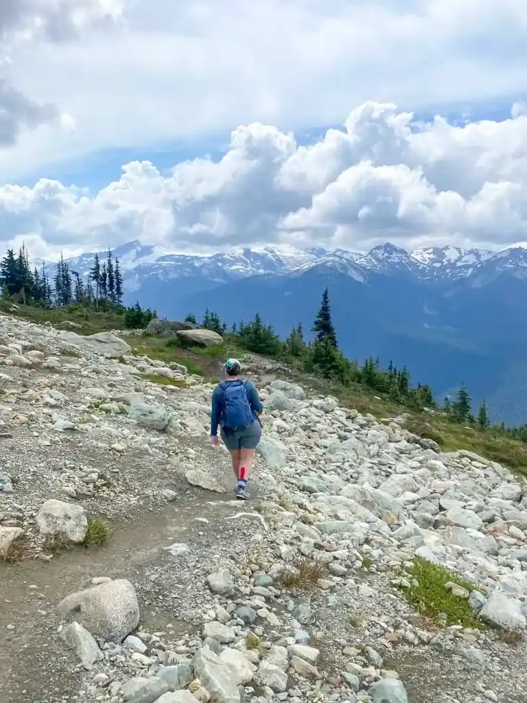 A female hiker wearing a blue backpack walks along a rocky trail with snow capped mountains in the background.