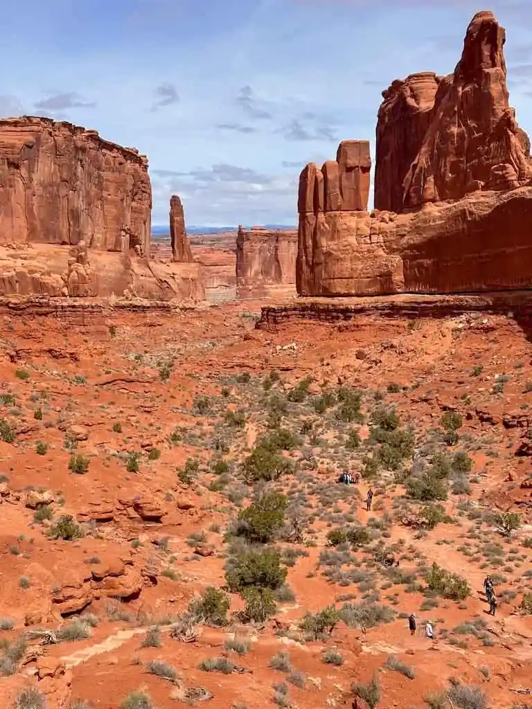 Looking north along the Park Avenue Trail in Arches National Park. There is a trail with hiker in the foreground. The trail passes between tall red rock spires.