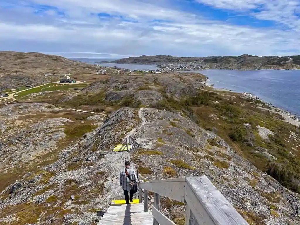 A man walks down wooden stairs on the way down from the viewpoint at Brimstone Head on Fogo Island, Newfoundland. The town of Fogo is visible in the distance.