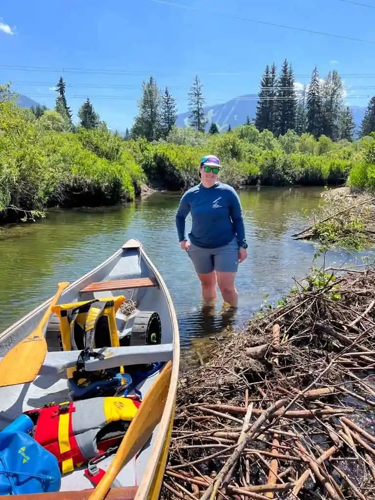 A woman stands in a river next to a beaver dam and a canoe. She is wearing the Mountain Hardwear Crater Lake Hoody