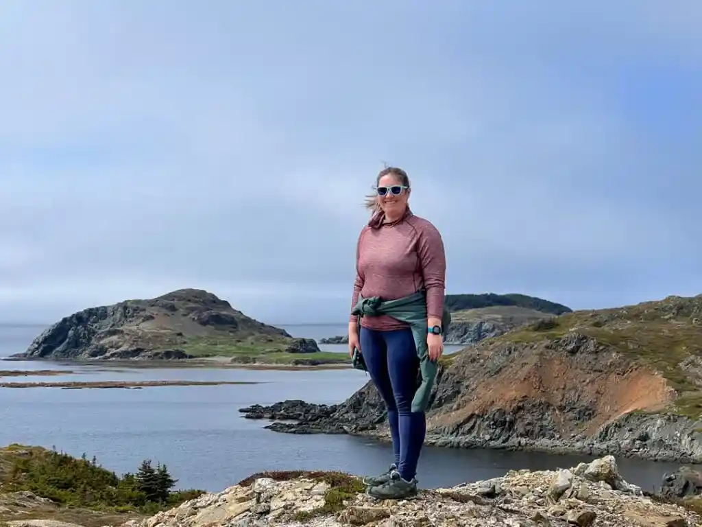 A woman poses on a hike in Twillingate. She is wearing a warm jacket tied around her waist.