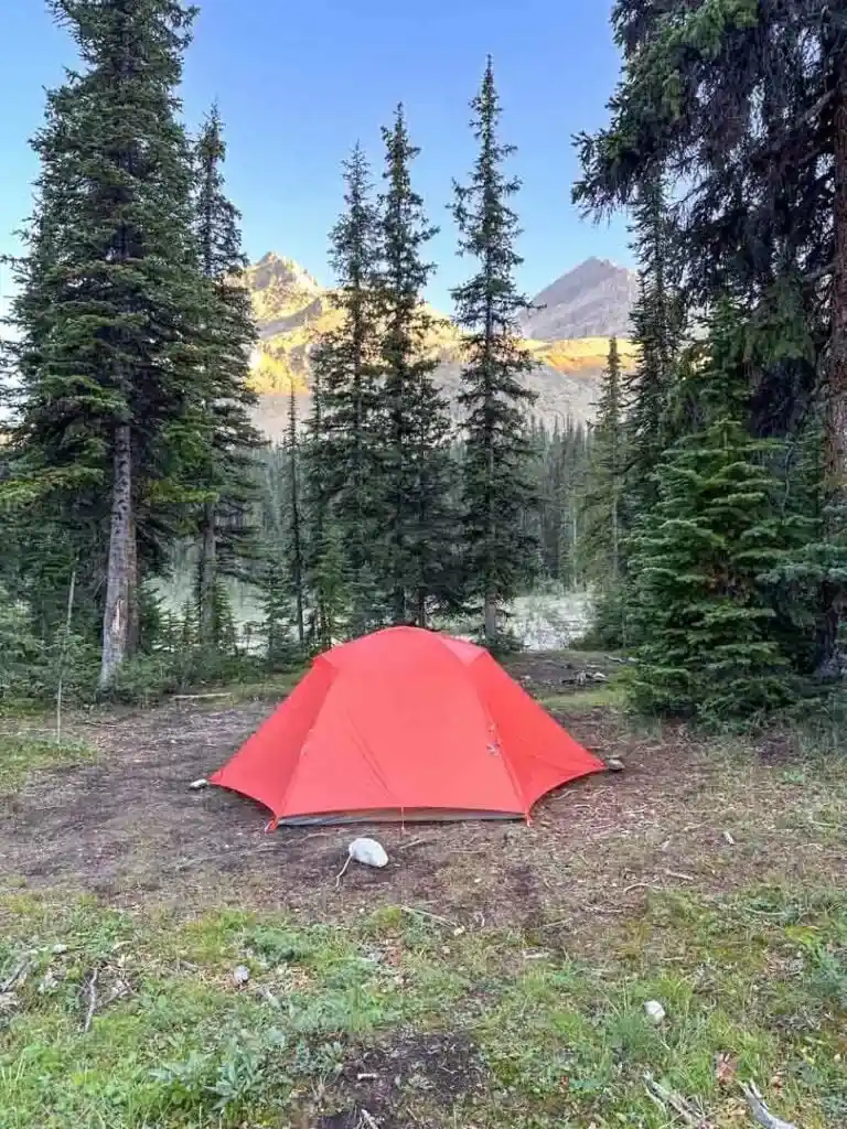 An orange Big Agnes Copper Spur tent at Little Yoho Camp in the Yoho National Park backcountry. You can see sun-dappled mountains in the background across a river.